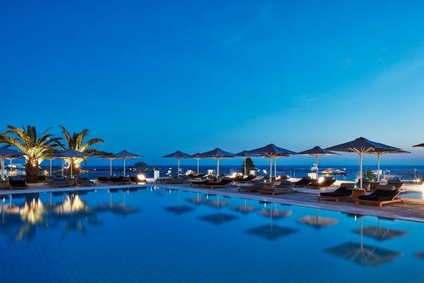 Luxurious white-walled Scenset Ambassador Hotel overlooking Elia Beach, with infinity pool, elegant sunbeds, and panoramic Aegean Sea views in Mykonos, Greece