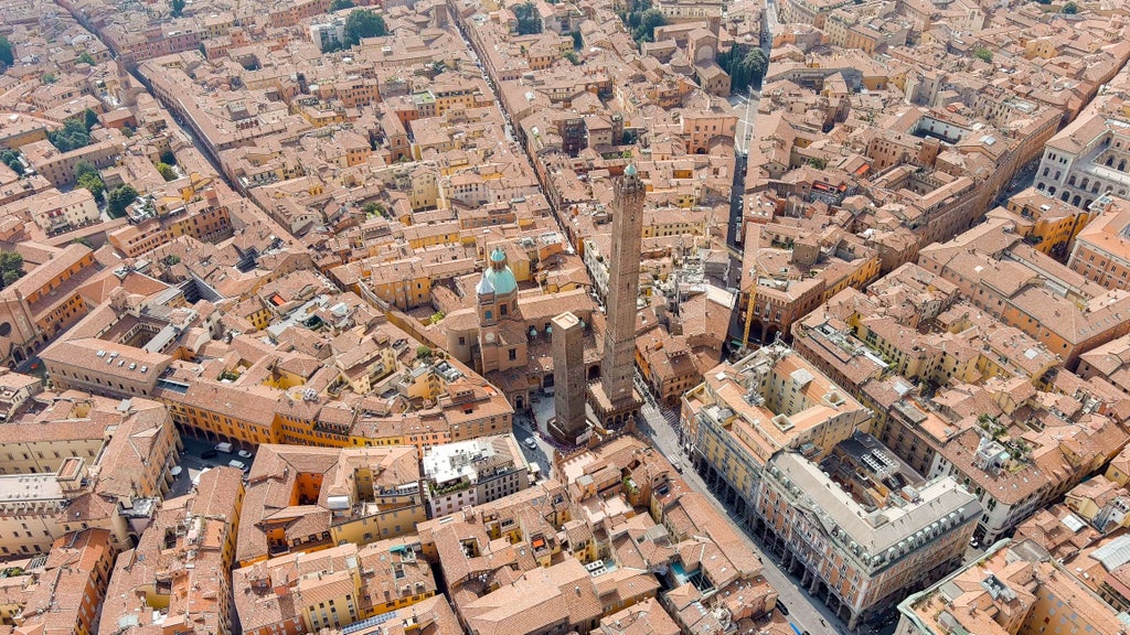 Medieval red-brick towers and elegant porticoes line Bologna's historic center plaza, bathed in warm evening light beneath clear Italian skies