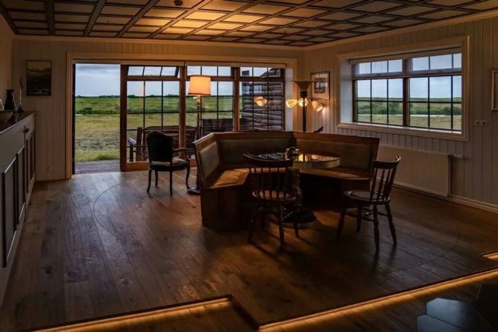 Luxurious master suite with king bed, chandelier, and panoramic windows overlooking Iceland's wilderness. Wood-paneled walls add warmth.