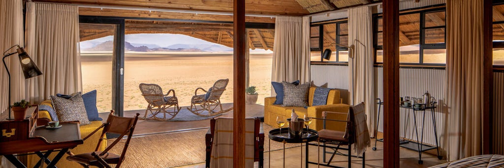 Modern luxury safari lodge with thatched-roof chalets nestled among red sand dunes of Namibia's desert, panoramic glass walls reflect sunset