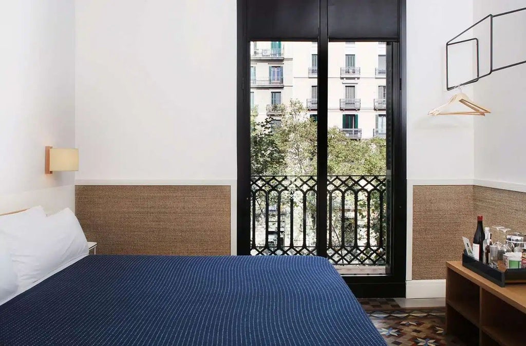 Modern, minimalist hotel room with warm wooden furnishings, crisp white linens, and large windows overlooking Barcelona's vibrant Gran Via streetscape.