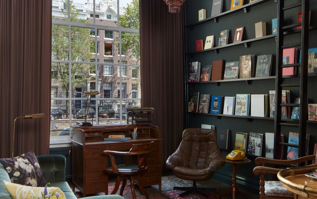 Elegant dark wood-paneled suite with floor-to-ceiling bookshelves, plush velvet sofa, ornate chandelier and large windows overlooking Amsterdam