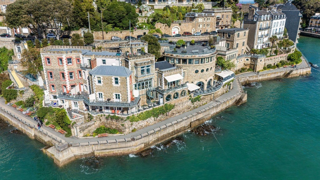 Luxurious coastal hotel with blue shutters and white facade, perched on rocky cliffs overlooking the azure waters of a French seaside town.