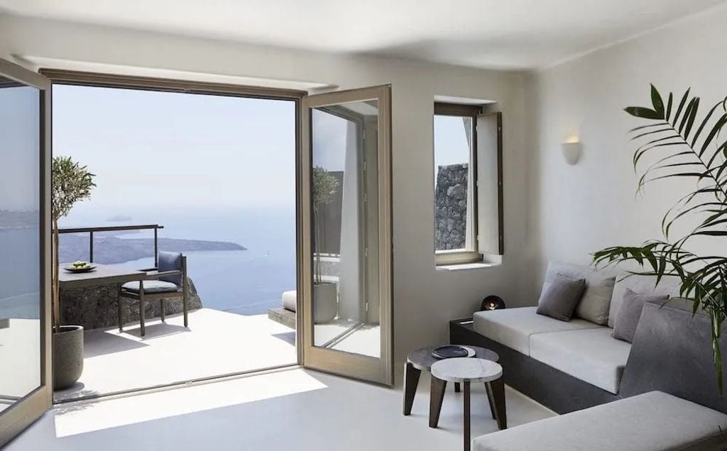 White Cycladic cave hotel suite with private infinity pool carved into Santorini cliffs, overlooking the Aegean Sea and caldera at sunset