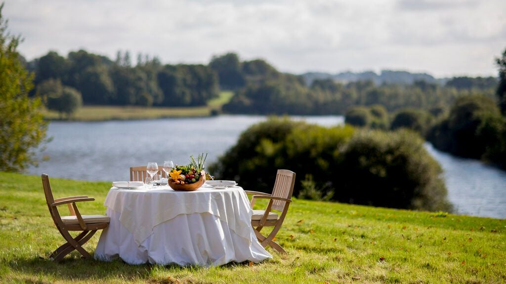 Luxurious stone castle-hotel nestled in lush French countryside, featuring elegant architecture, expansive green grounds, and serene pond reflections at twilight