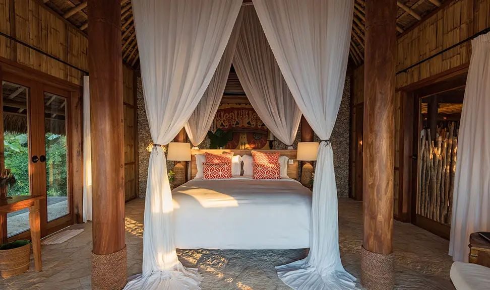 Luxurious open-air villa with sweeping views of lush Indonesian landscape, featuring thatched roof, infinity pool, and elegant wooden furnishings at NIHI Sumba resort