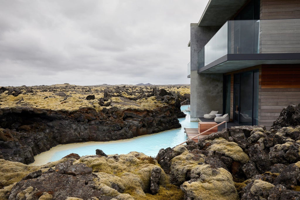 Luxurious modern hotel nestled in dramatic Icelandic landscape, featuring minimalist design with floor-to-ceiling windows overlooking stark volcanic terrain and distant mountains.