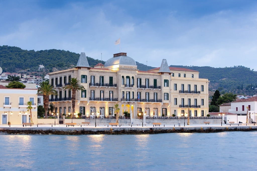 Elegant neoclassical waterfront hotel with white facade, grand columns, and palm trees overlooking the Mediterranean Sea at sunset