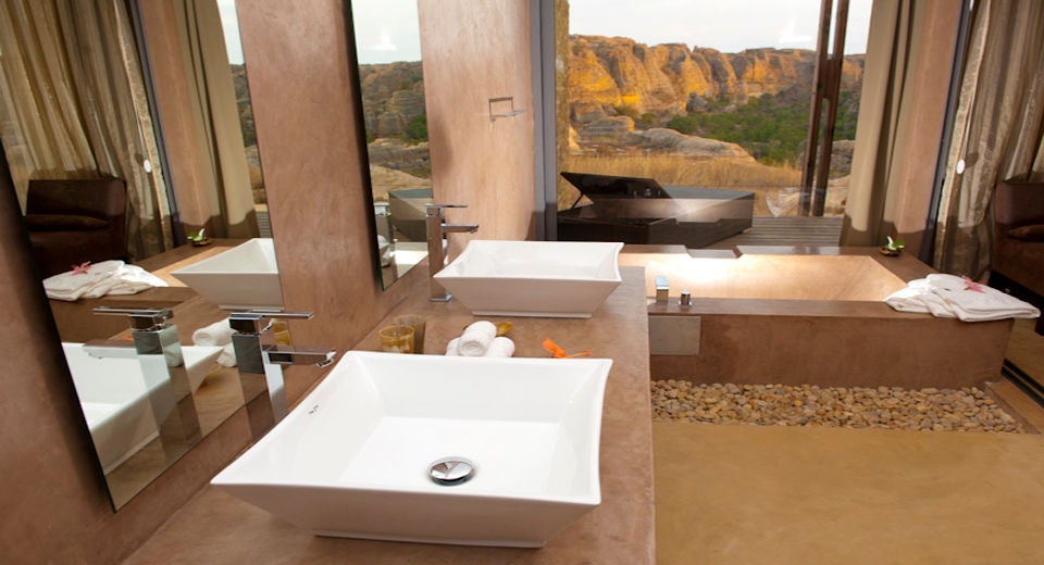 Modern luxury lodge perched on rocky cliffs in Madagascar, featuring glass-fronted suites overlooking vast Isalo sandstone formations at sunset