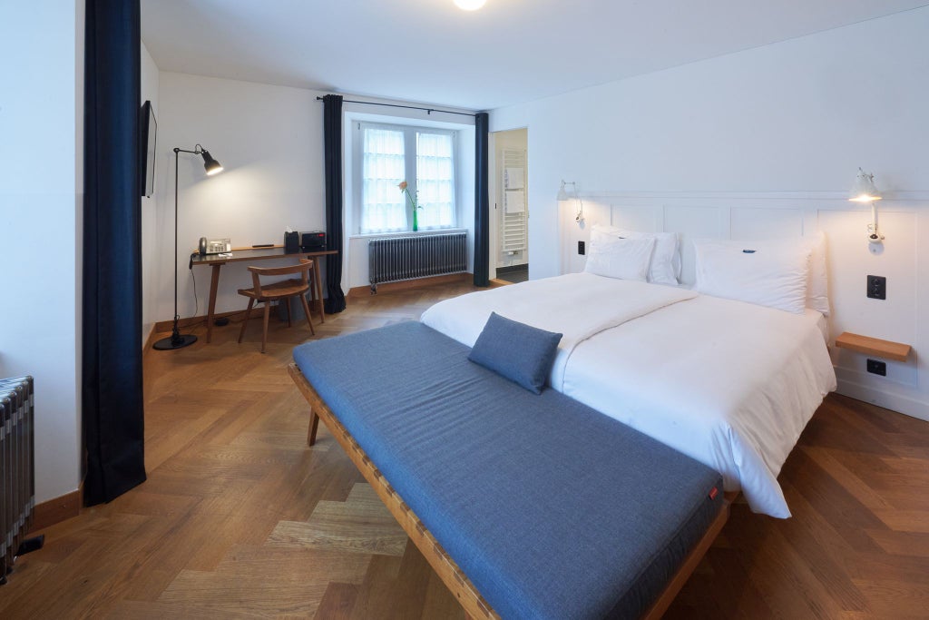 Elegant Swiss hotel room with plush white bedding, modern minimalist decor, large windows, and soft neutral color palette overlooking urban scenery