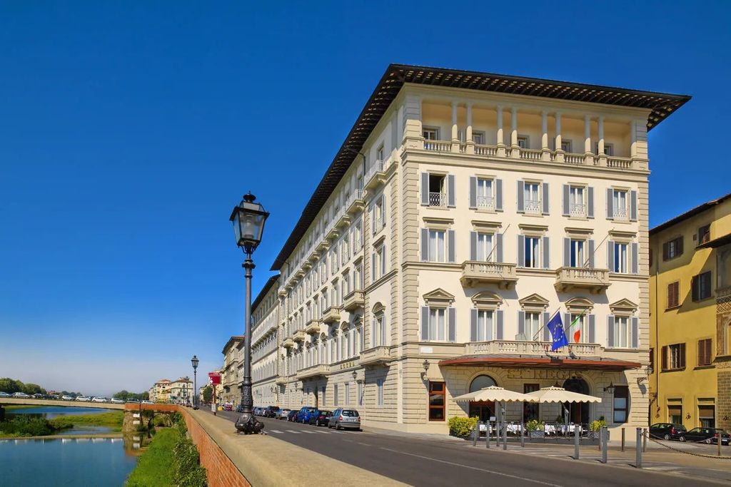 Opulent Renaissance-style hotel facade with ornate stone carvings, arched windows and wrought iron balconies overlooking Arno River in Florence