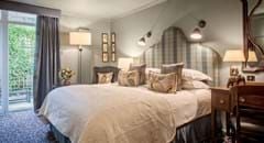 Classic English hotel room with elegant neutral tones, plush white bedding, traditional wooden furniture, and soft ambient lighting in a charming Cotswolds-style setting