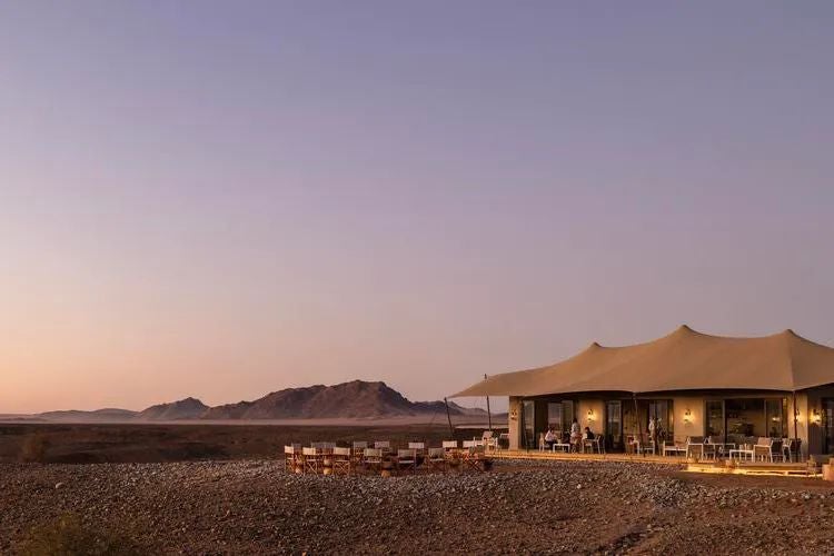 Luxurious eco-lodge nestled in rugged Namibian desert landscape, with rustic wooden cabins overlooking dramatic rocky terrain and sparse, golden vegetation.