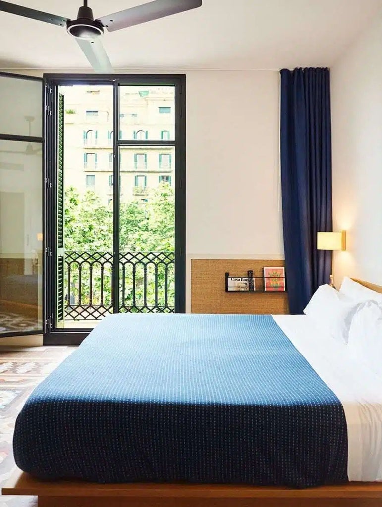 Minimalist chic hotel room with elegant white bedding, wooden accents, and large windows overlooking Barcelona's urban landscape at Casa Bonay.