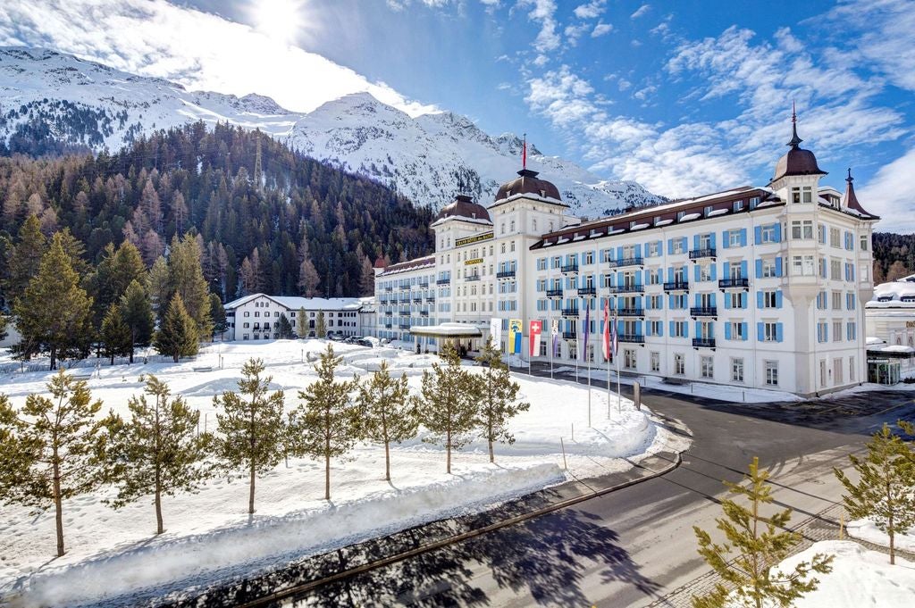 Luxurious alpine resort featuring white facade with ornate balconies, surrounded by snow-capped mountains and manicured gardens