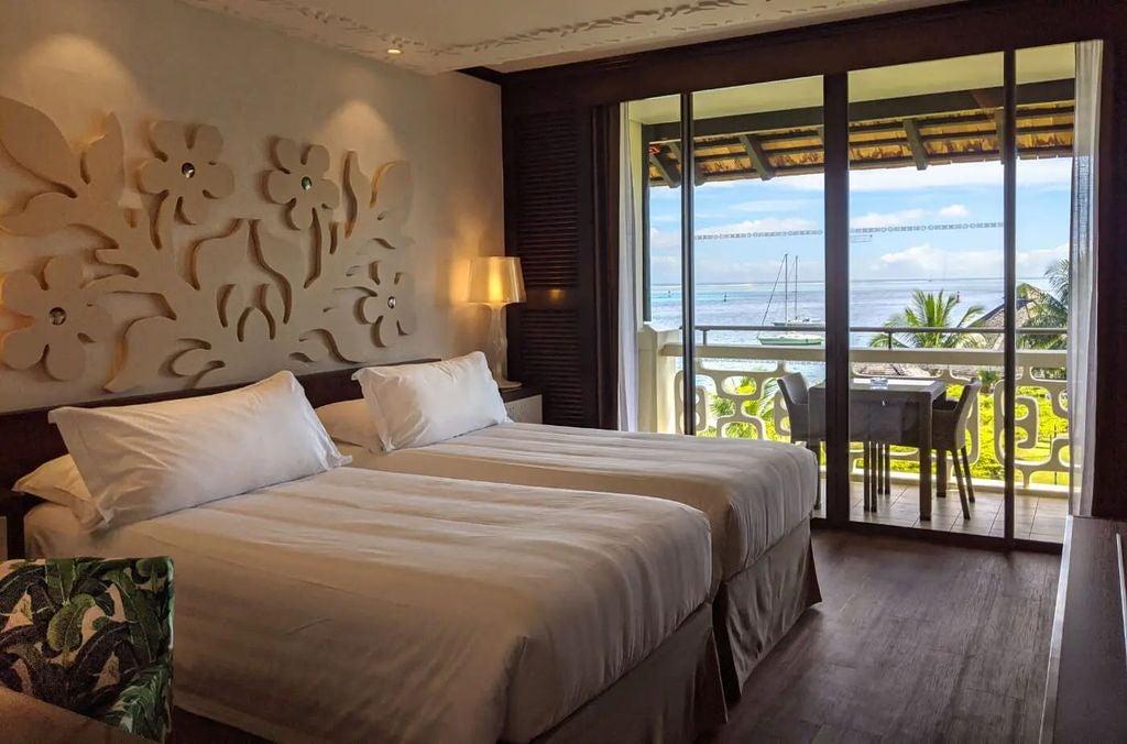 Luxurious hotel room with king bed, white linens, dark wood furniture, tropical decor, and floor-to-ceiling windows overlooking ocean views