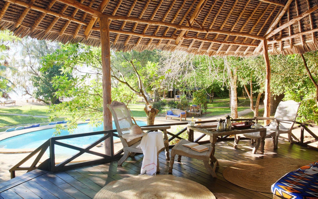 Tropical beachfront cottage with white walls, thatched roof, wooden deck, swaying palm trees, and turquoise Indian Ocean view in coastal Kenya.