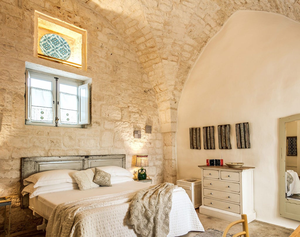 Elegant rustic Italian hotel room with stone walls, wooden furnishings, white linens, and a traditional arched window overlooking scenic countryside landscape