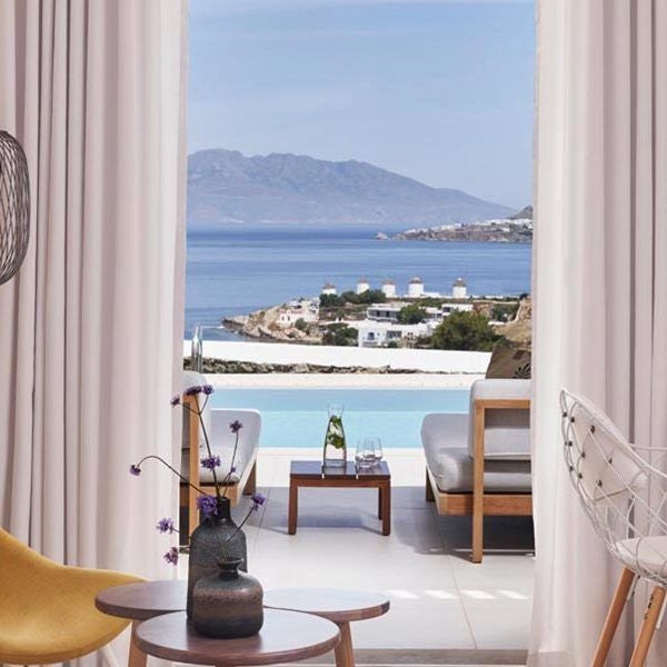 Luxurious white-walled Horizon Superior Room at Myconian Kyma, featuring minimalist design, blue-tinted windows overlooking Aegean Sea with crisp, elegant bedding