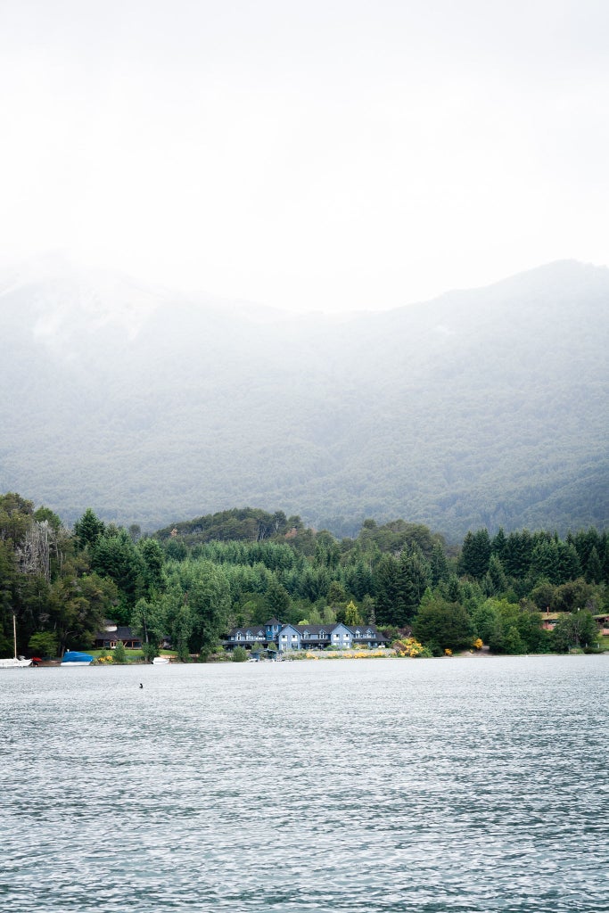 Lakeside luxury lodge with wooden deck overlooking Patagonian mountains, featuring elegant outdoor seating and natural stone accents