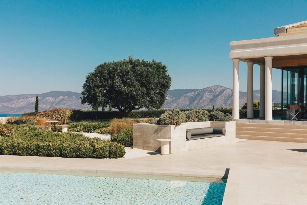 Modern luxury resort with white stone columns and infinity pool overlooking Aegean Sea and rolling Greek coastal hills at sunset