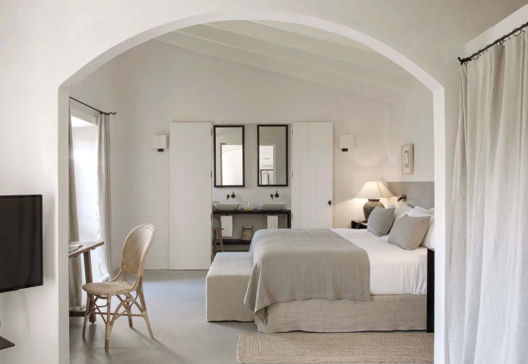 Elegant suite with four-poster bed, white linens and rustic wooden beams overhead, offering views of Mallorca's countryside through arched windows