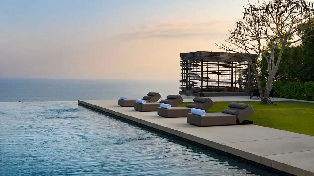 Infinity pool overlooking Indian Ocean cliffs at sunset, modern villa with wooden deck and white loungers in tropical setting