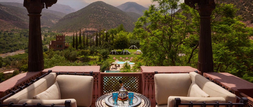 Traditional Moroccan hilltop hotel with red clay walls, blue pool, and lush gardens against dramatic Atlas Mountain backdrop at sunset