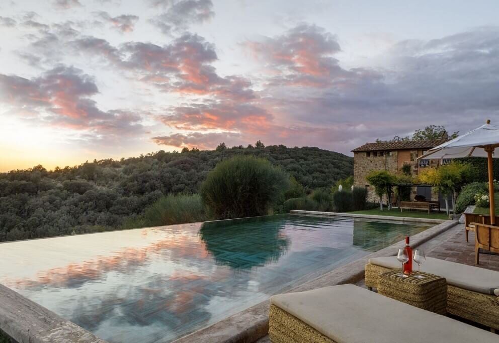 Luxurious stone Tuscan castle hotel nestled among rolling olive groves, with sunset-lit terracotta roof and elegant stone facade against verdant landscape