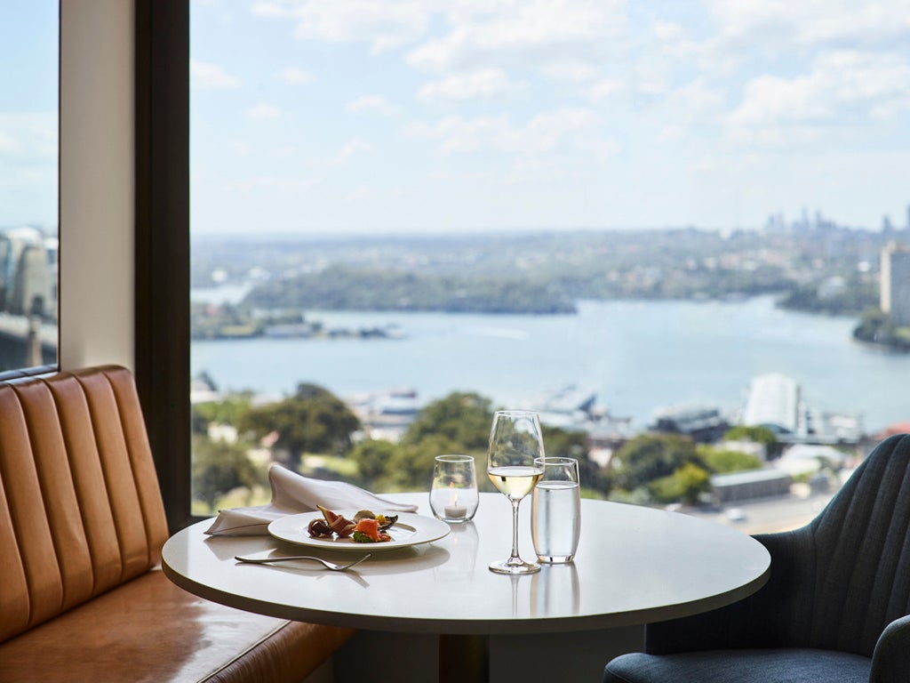 Modern luxury high-rise hotel with glass facade overlooking Sydney Harbour Bridge, illuminated at dusk with warm city lights