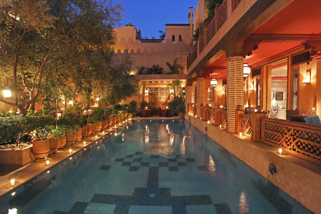 Traditional Moroccan riad with ornate courtyard featuring mosaic tiles, stone fountain, tropical plants and luxurious poolside loungers at dusk