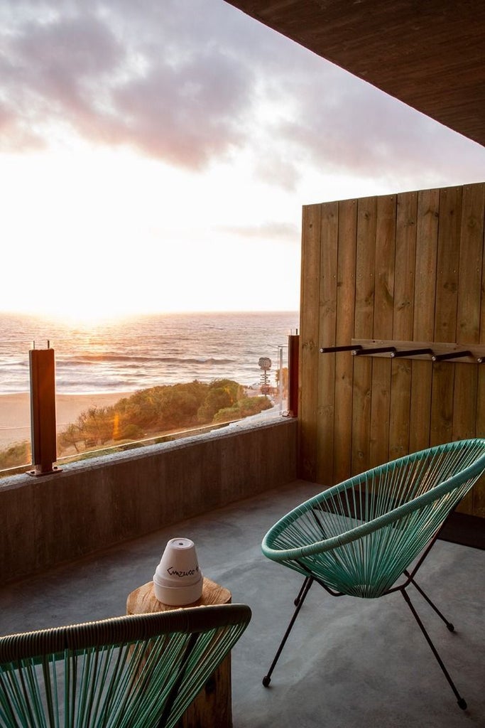 Minimalist surf-inspired hotel room with ocean view, white linens, wooden accents, and floor-to-ceiling windows overlooking coastal landscape in Portugal