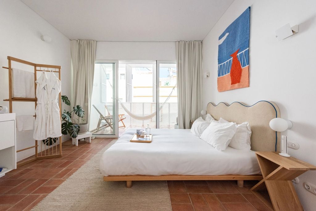 Clean, minimalist hotel room with rustic white walls, wooden bed frame, woven rug, and natural textiles bathed in soft sunlight through arched windows.