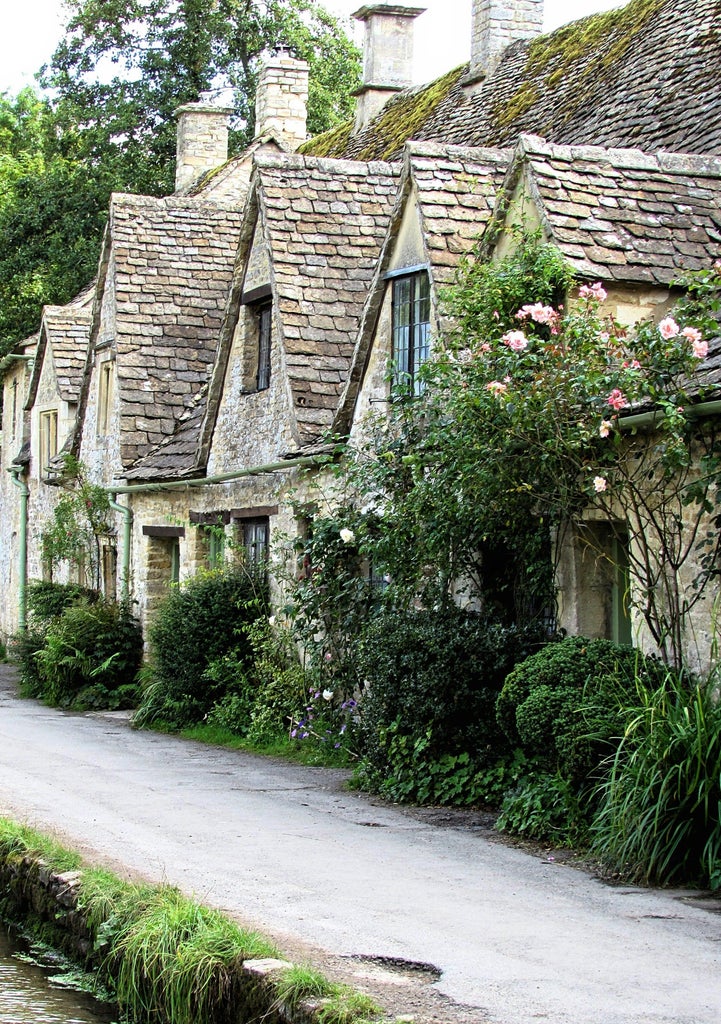 Rolling green hills dotted with honey-colored stone cottages and blooming gardens in a historic Cotswolds village under soft clouds