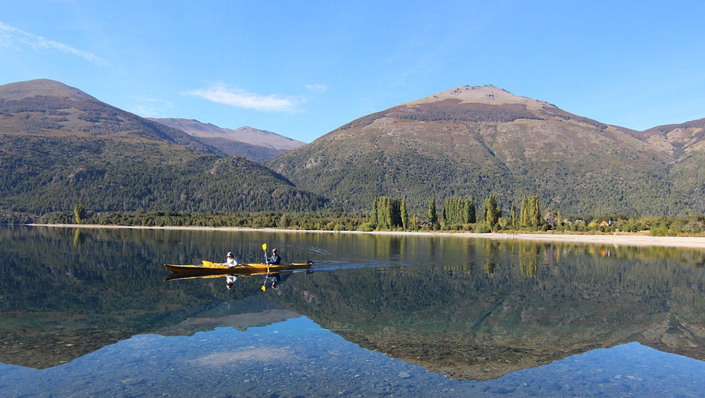 Luxurious mountain lodge with wooden facade nestled against snow-capped Andes peaks, overlooking serene Nahuel Huapi lake at sunset