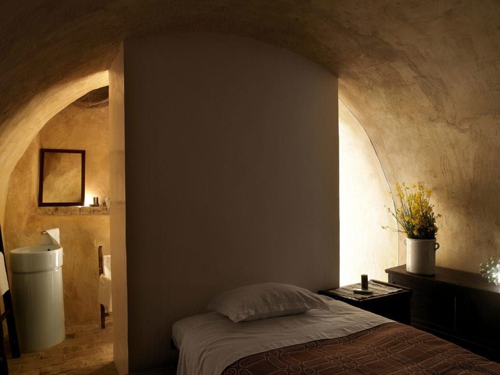 Rustic stone-walled Italian hotel room with antique wooden furniture, soft linen bedding, and warm terracotta floor tiles in soft afternoon light