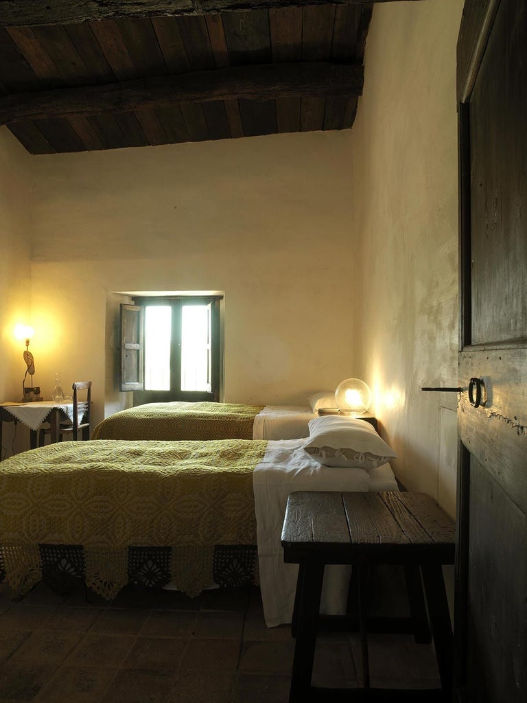 Stone-walled rustic Italian hotel room with exposed wooden beams, antique furnishings, and soft natural light filtering through arched window overlooking medieval village landscape