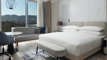 Elegant king room with floor-to-ceiling windows, modern furnishings, plush bedding, and sweeping city views of downtown Tirana