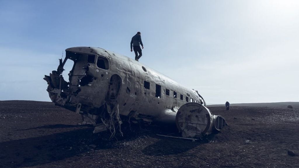 All-terrain vehicle traversing black volcanic sand near Iceland's famous crashed airplane wreckage, dramatic landscape with rugged terrain and volcanic mountains in background