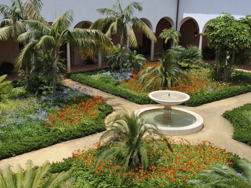 Elegant Mediterranean hotel with white-washed walls, arched walkways and manicured gardens, framed by mountains and palm trees