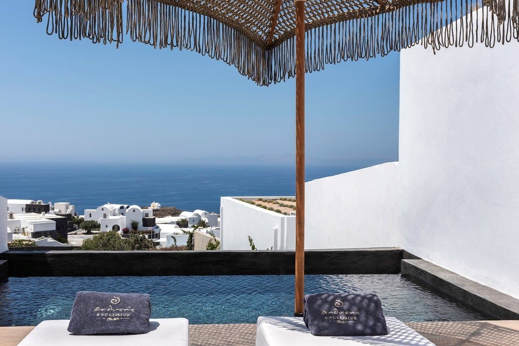 Two-bedroom luxury suite balcony with infinity pool overlooking Santorini's caldera, featuring elegant outdoor loungers and sea views