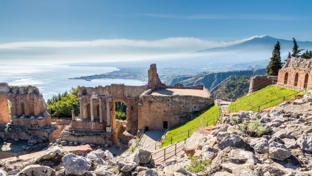 Panoramic view of Taormina's medieval town perched on cliffs, overlooking turquoise Mediterranean Sea with Mount Etna in background