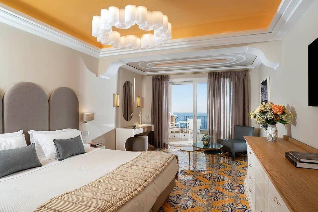 Elegant Italian hotel room with ornate gold-framed mirror, plush king-sized bed, crystal chandelier, and panoramic scenic window overlooking coastal landscape