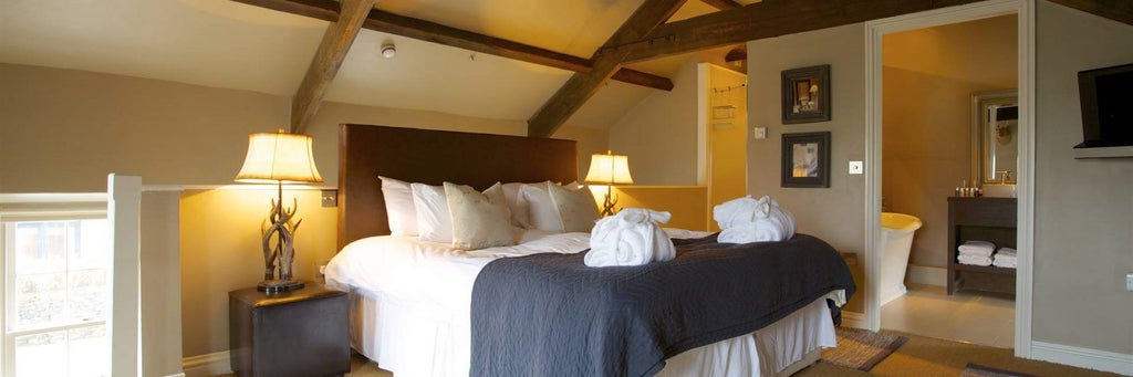 Elegant boutique hotel room with plush white bedding, soft neutral tones, contemporary furnishings, and expansive window overlooking scenic countryside landscape