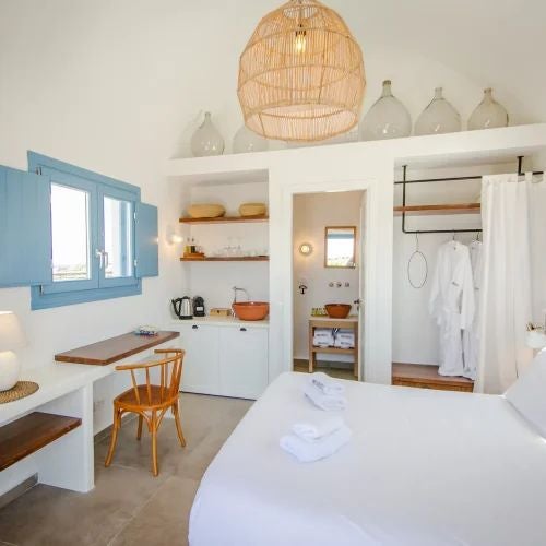 Elegant Mediterranean villa bedroom with wooden floors, white linens, and expansive terrace overlooking scenic Greek landscape with warm afternoon sunlight