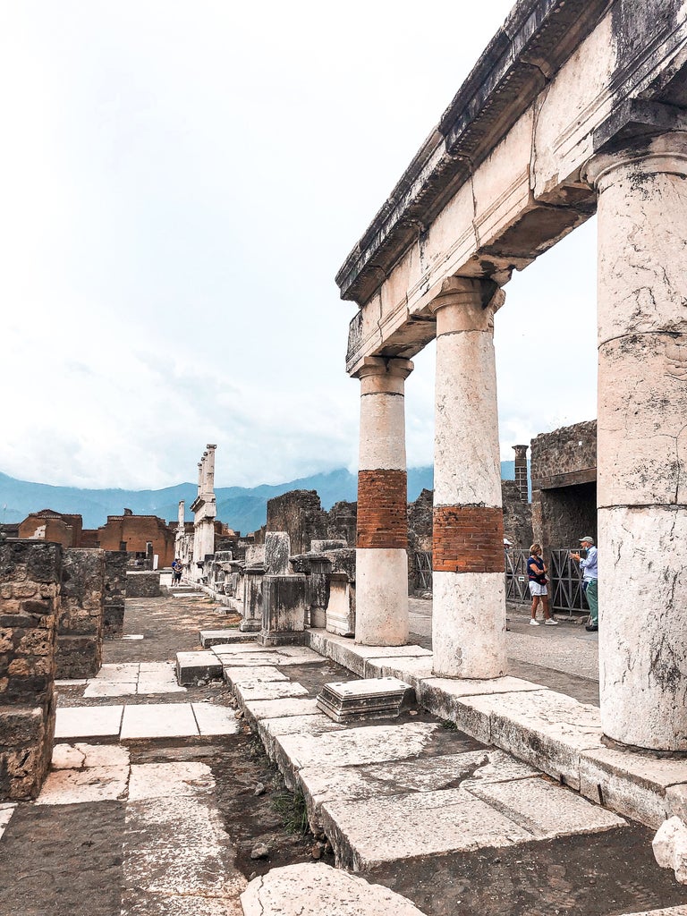 Ancient stone columns and archways of Pompeii ruins against Mount Vesuvius, bathed in warm Mediterranean sunlight at golden hour