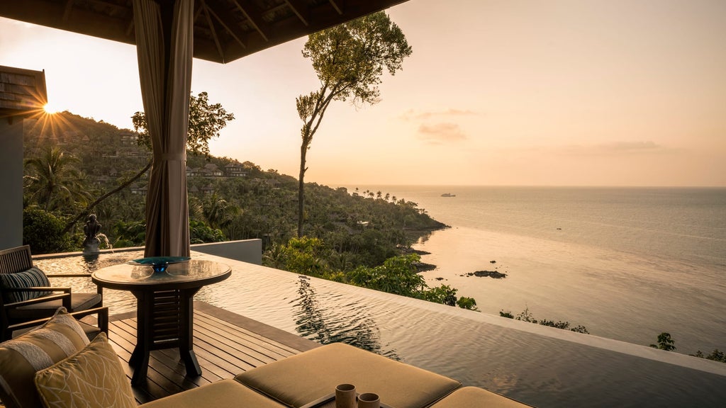 Infinity pool with white lounge chairs overlooking turquoise ocean and lush tropical hillside at sunset, nestled in Koh Samui's coastline