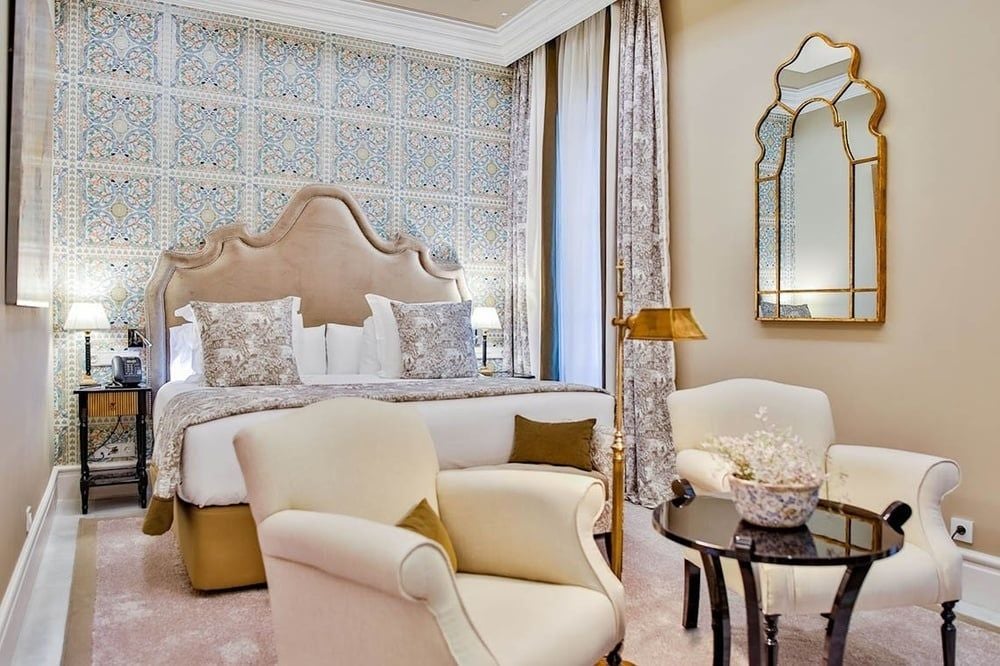 Elegant deluxe hotel room with crystal chandelier, ornate gold mirror, velvet armchair, and period furniture against cream-colored walls