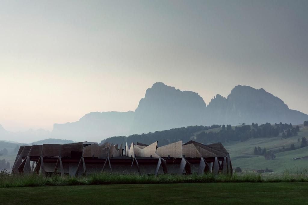 Modern alpine resort with panoramic glass windows and wooden facade nestled against dramatic Dolomite mountains in snowy landscape