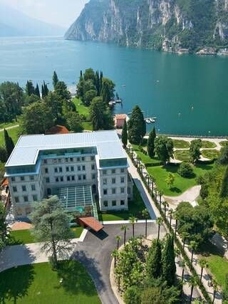 Elegant Art Nouveau hotel facade with ornate white balconies overlooking Lake Garda, surrounded by manicured gardens and alpine mountain backdrop